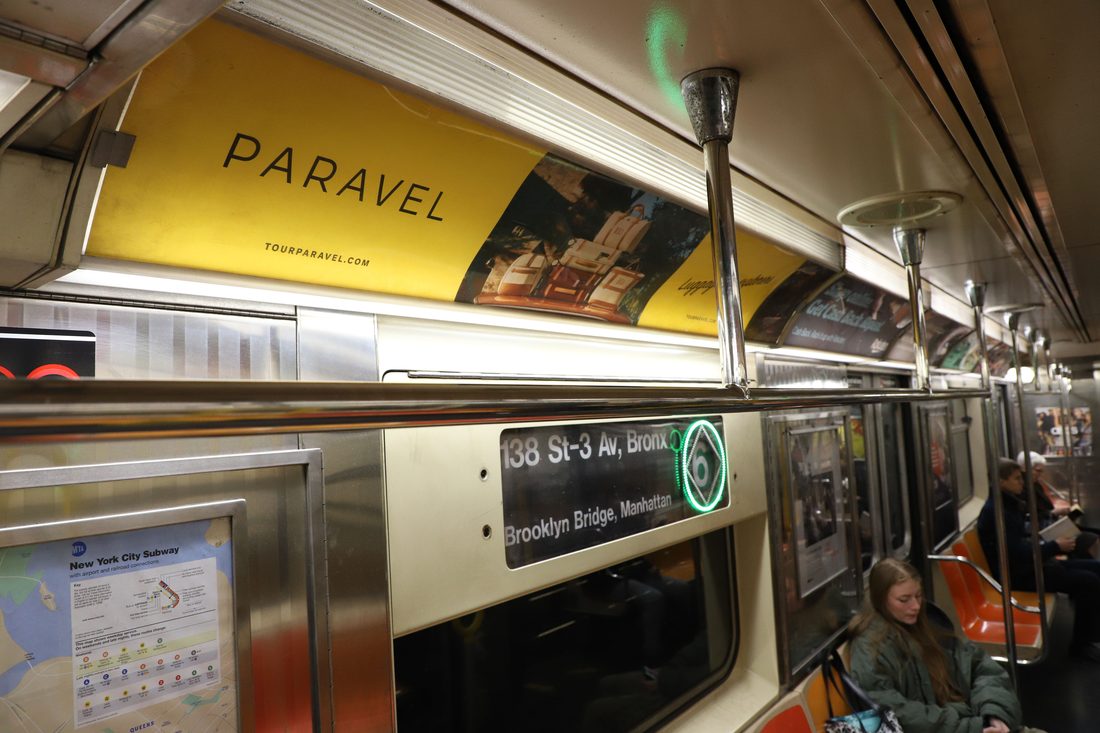 Inside an NYC subway car: a yellow PARAVEL ad panel of luggage runs above the windows, beside the train's route sign.
